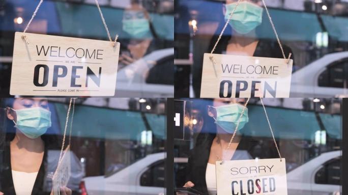 企业主迷人的亚洲年轻人围裙挂着我们前门的开放标志欢迎客户来到新咖啡馆。站在咖啡馆时，戴着防护面罩的快