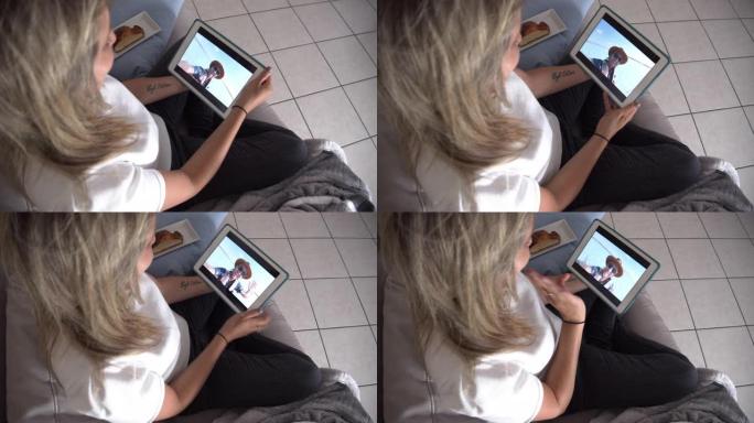 年轻女子在家使用数字平板电脑与男友交谈时在沙发上放松的后视图。男子通过笔记本电脑上的视频聊天进行交流