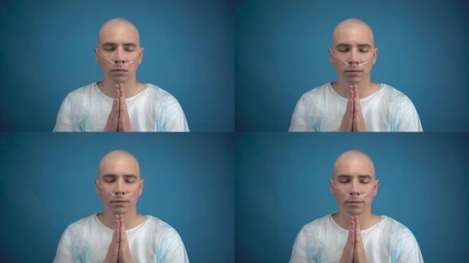 秃头的年轻人和肿瘤学人看着相机，在蓝色背景上祈祷。病人双手合十，微笑着祈祷。化疗导致脱发。
