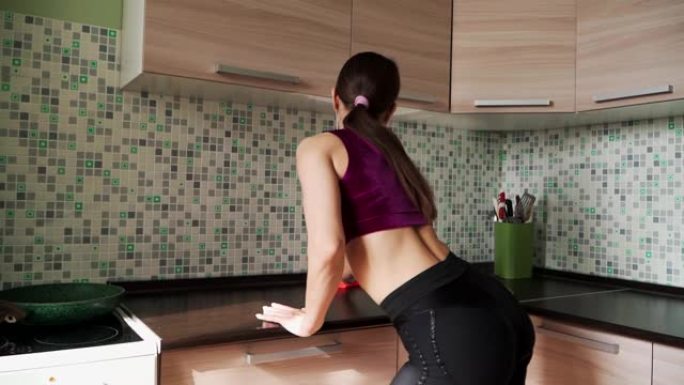 女人性感的运动战利品穿着黑色紧身紧身裤戴着耳机在厨房里用抹布跳舞，擦家具。慢动作