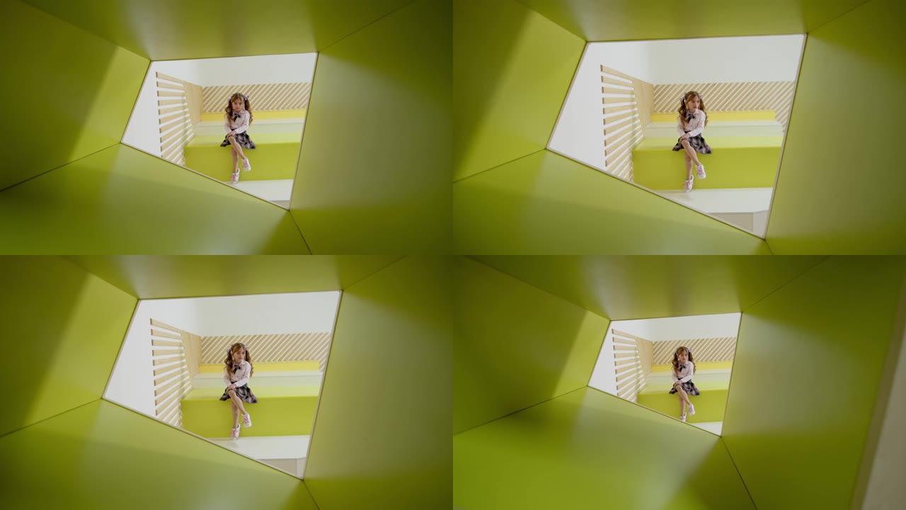 一个小女孩独自坐在黄色和绿色的抽象彩色长凳上，看着相机。摄像机穿过一个木洞