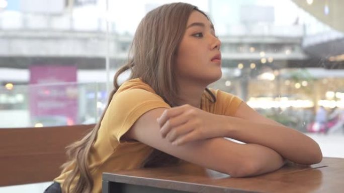 坐在咖啡馆窗边的沮丧年轻美丽的亚洲女人的高清慢动作。孤独悲伤的女孩从窗外望着城市街道，思考问题。年轻