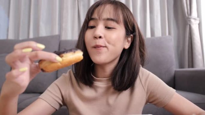 年轻的亚洲女性外卖在客厅吃甜甜圈垃圾食品在家里享受幸福，快餐送货服务安全，并通过社交距离概念阻止冠状