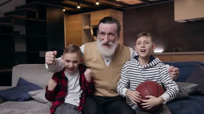 令人兴奋的正面两个十几岁的男孩和他们受人尊敬的大胡子祖父，他们在家里的电视上一起看体育比赛