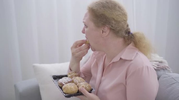 沮丧的胖乎乎的女人嚼着甜面包，哭了起来。紧张的高加索高级女士抑郁症的侧视图。悲伤，垃圾食品，压力饮食