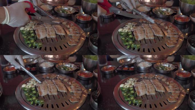 在韩国餐厅，厨师手用食物剪剪烟熏烤架猪肚肉在韩国铁烧烤烤盘上拍摄4k特写镜头。传统的韩式食品烧烤猪肚