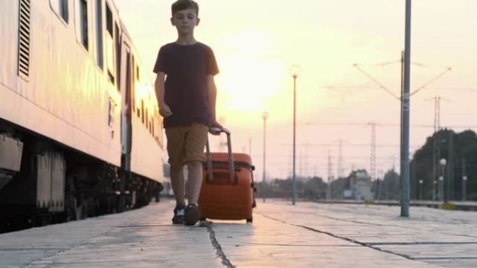 快乐男孩橙色手提箱在明亮阳光明媚的日落慢动作中沿着他身后火车站的平台滚动行走。孩子坐火车去度假。旅游