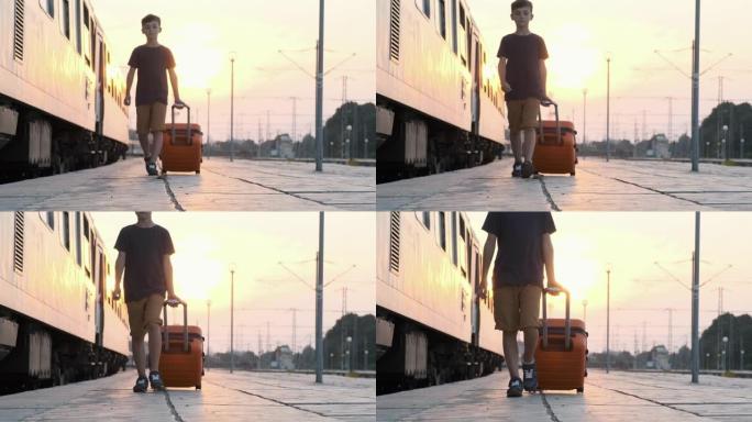 快乐男孩橙色手提箱在明亮阳光明媚的日落慢动作中沿着他身后火车站的平台滚动行走。孩子坐火车去度假。旅游