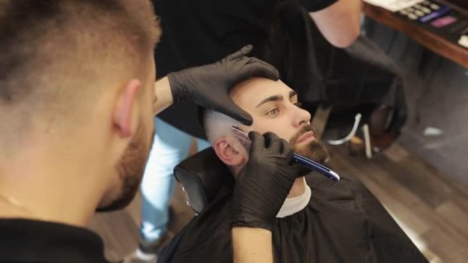 戴着黑色手套的手刮男人的脸。专业理发师用直剃刀刮顾客胡须。理发店用老式刀片切割胡须