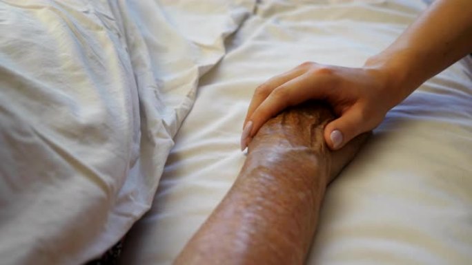 孙女在医疗诊所握住并安慰祖母的手。女孩轻轻地抚摸躺在医院病床的生病的成熟女人的皱纹手臂。女儿对母亲表