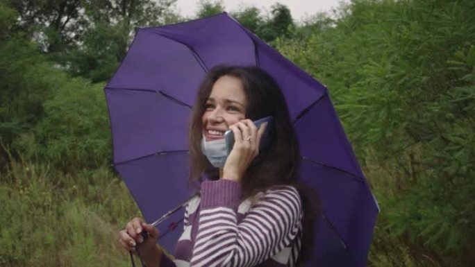 在阴天的秋天森林里，戴着冠状病毒面具的年轻白人妇女拿着雨伞，用智能手机。