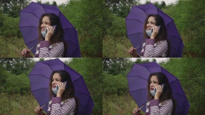 在阴天的秋天森林里，戴着冠状病毒面具的年轻白人妇女拿着雨伞，用智能手机。