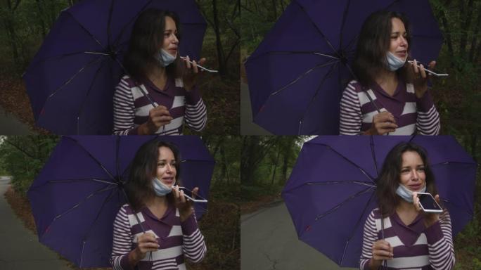 在阴天的秋天森林里，戴着冠状病毒面具的年轻白人妇女拿着雨伞，用智能手机。一名妇女在秋天的森林中的智能