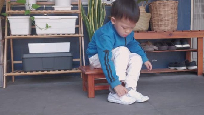 亚洲3岁幼稚园孩子坐在靠近他家前门的鞋架，集中精力穿上他的白色鞋子/运动鞋