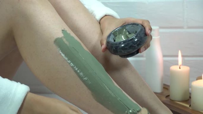 一名妇女用抹刀将粘土面膜涂在腿部皮肤上。脱毛后的刺激，剃须。家庭护肤，身体。水疗，保湿，治疗。去角质