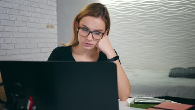 疲倦困倦的高加索年轻女子在家里用笔记本电脑工作时打哈欠并入睡。戴眼镜的金发女孩晚上在笔记本电脑上工作