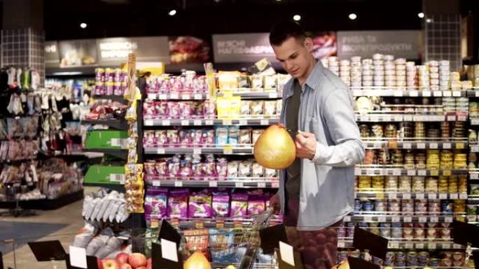 一个微笑的白人男子，穿着蓝色衬衫，在一家杂货店超市的生产部门购买水果和蔬菜。带着柚子，把它放进购物车