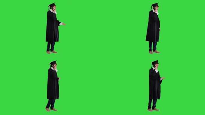 穿着长袍和砂浆板的男性毕业生在绿屏上通过电话交谈，色度键