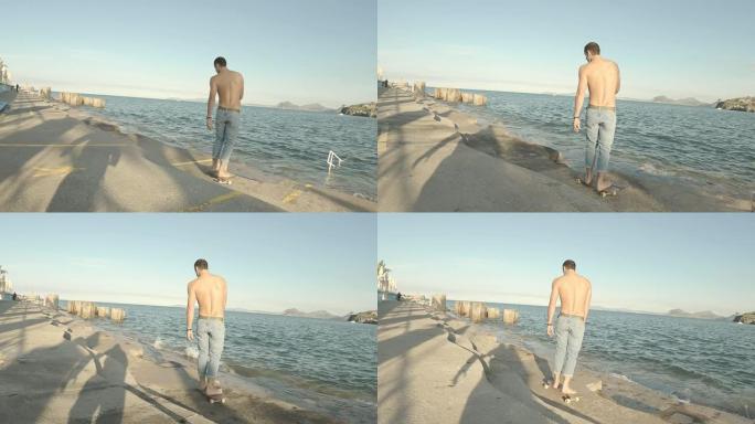 穿着蓝色牛仔裤的年轻人在下午以大海为背景的码头上赤脚光着膀子溜冰
