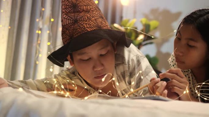 亚洲儿童戴着巫婆的帽子躺在床上，晚上在家用led灯读童话书，万圣节概念。