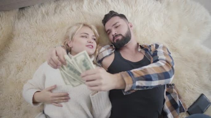 幸福富有的高加索夫妇躺在柔软的地毯上数钱。穿着白色毛衣的金发女孩和英俊的大胡子男人在家休息。财富、幸