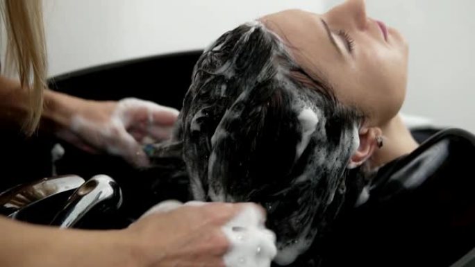 美容工作室。用洗发水洗深色长发的女人。按摩头部雕塑。收起理发师的手在头上洗头，起泡沫。放松水疗程序