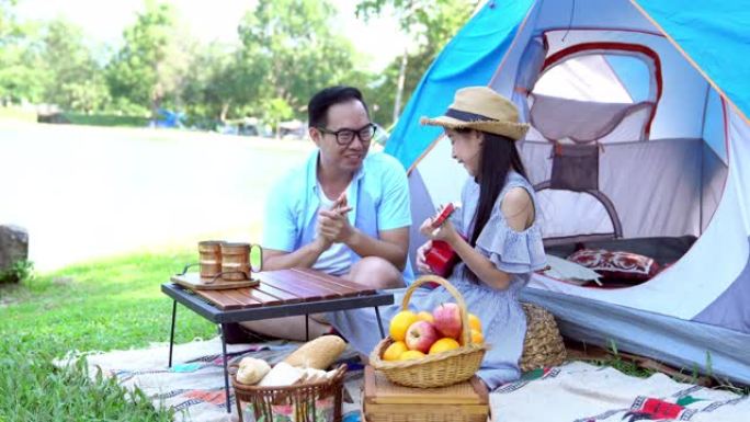 4k快乐的亚洲男人父亲和漂亮的女孩女儿放松身心，享受夏天在湖边露营的户外活动，一起演奏尤克里里吉他。