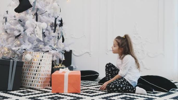 快乐的孩子在圣诞节早上来到枞树，想拿一份用红纸包裹的礼物，但是里面确实有些东西很重，所以女孩无法捡起