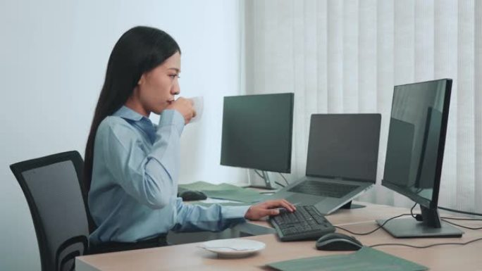 亚洲妇女在清晨喝咖啡，同时考虑计算机屏幕上的程序和代码。