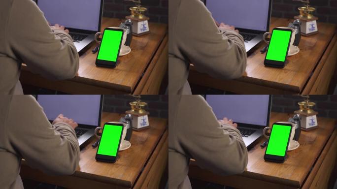 带有绿色屏幕的模型，用于跟踪特写。成年男子坐在桌子旁，在笔记本电脑上输入文字。附近有一部带触摸屏和老