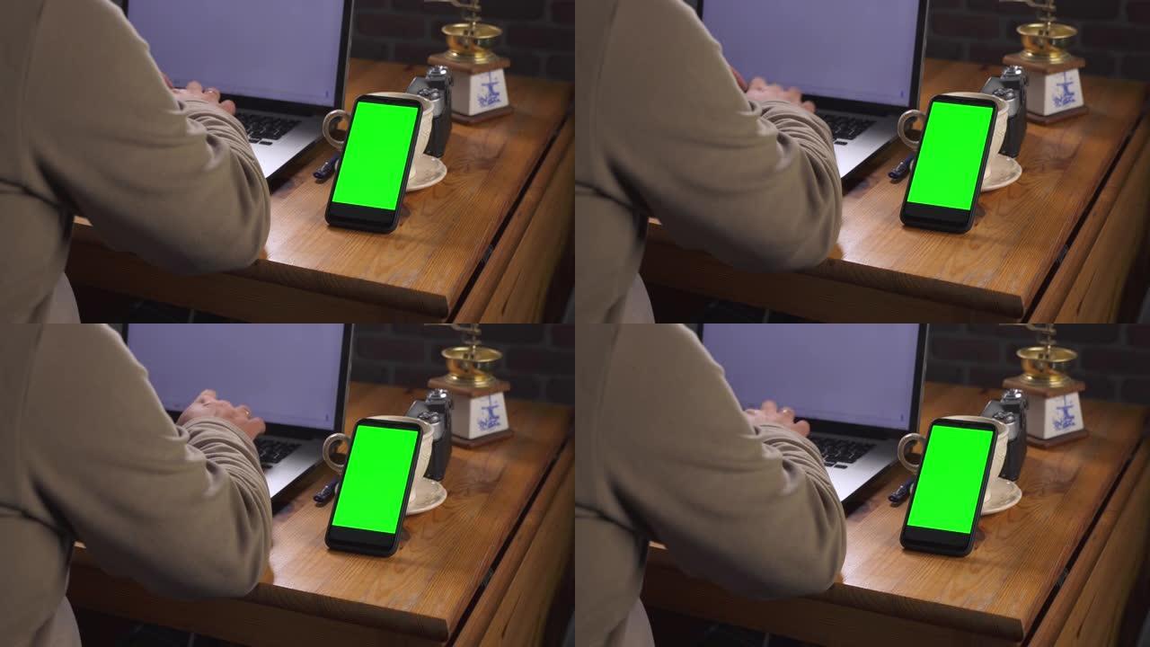 带有绿色屏幕的模型，用于跟踪特写。成年男子坐在桌子旁，在笔记本电脑上输入文字。附近有一部带触摸屏和老