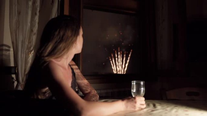 悲伤的女人独自一人坐在桌旁，手里拿着酒杯，透过家中的窗户观看令人惊叹的五颜六色的烟花。女性看着户外的