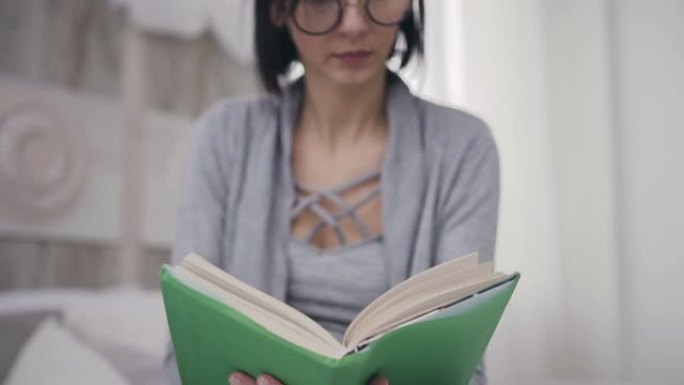 美丽的黑发女孩眼镜坐在床上，拿着一本书供阅读。一名年轻女子在家中用绿色封面打开一本有趣的书