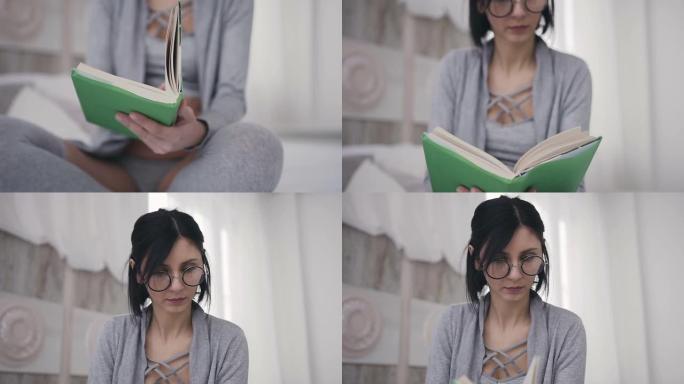 美丽的黑发女孩眼镜坐在床上，拿着一本书供阅读。一名年轻女子在家中用绿色封面打开一本有趣的书