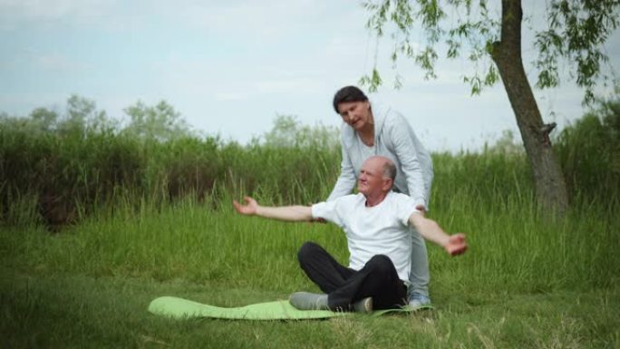 老年男女夫妇坐在绿色草地的瑜伽垫上进行健康锻炼，爱心妻子通过举起双手帮助丈夫锻炼身体