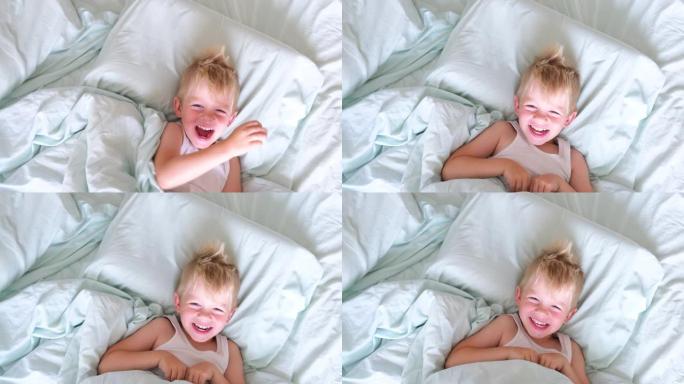 一个金发碧眼的小男孩躺在床上微笑着，玩得很开心，躲在被窝下，清晨从他下面偷窥。该醒来了