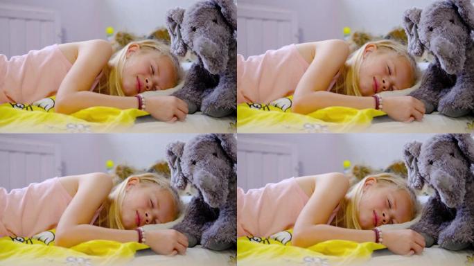 穿着粉色睡衣的漂亮金发小女孩一大早就睡在床上，穿着黄色床上用品。女孩和她的泰迪灰色大象