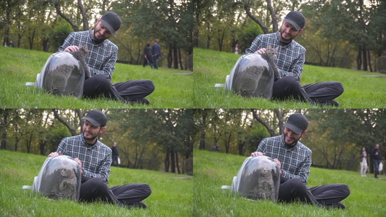 带宠物旅行的主题设备。带宠物徒步旅行的透明载体。男人和坐在公园绿草地上的灰猫玩耍。雄性使用呼吸胶囊袋