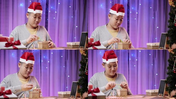 快乐的亚洲男孩戴着圣诞老人的帽子，在家用圣诞灯背景在礼品盒上写信息。