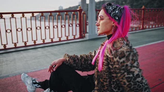 年轻的时髦女士，粉红色的头发，穿着非正式的衣服，侧身坐在沥青上，在桥上摆姿势