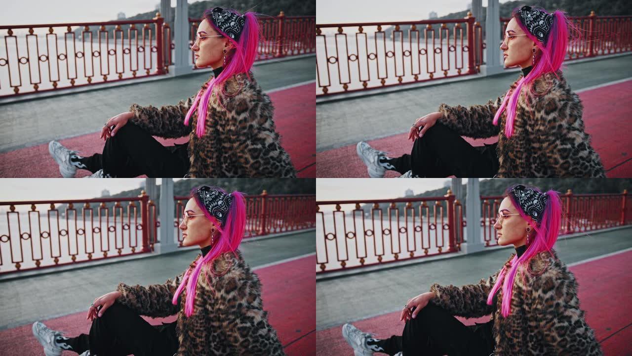 年轻的时髦女士，粉红色的头发，穿着非正式的衣服，侧身坐在沥青上，在桥上摆姿势
