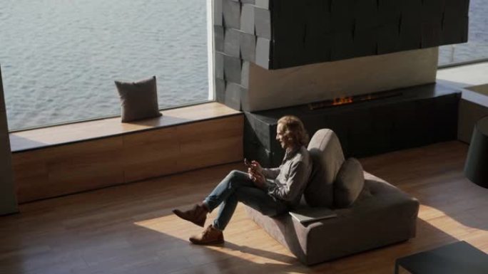 从上面拍摄的年轻人盘腿坐在他的豪华公寓的沙发上，他的大全景窗户可以看到海景和手机上的短信