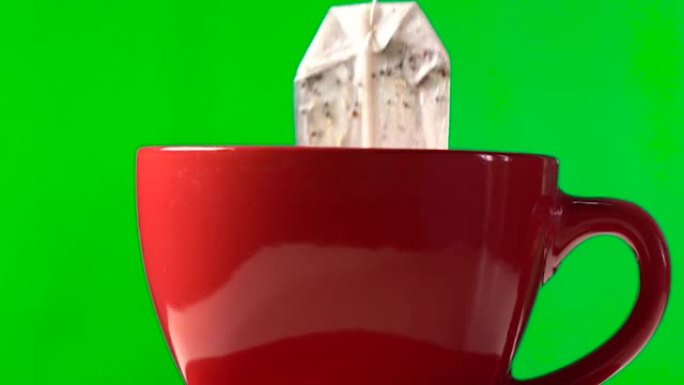 茶包在水里。孤立在绿色背景上。将茶包浸入带热水的红色杯子中，冲泡后，将茶包从杯子中取出