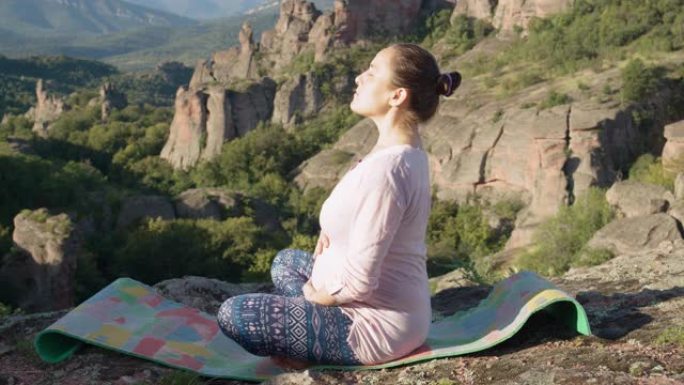 Vlogging教程。年轻的孕妇在户外做放松瑜伽练习，身后有雄伟的风景背景。展示如何在怀孕期间锻炼。