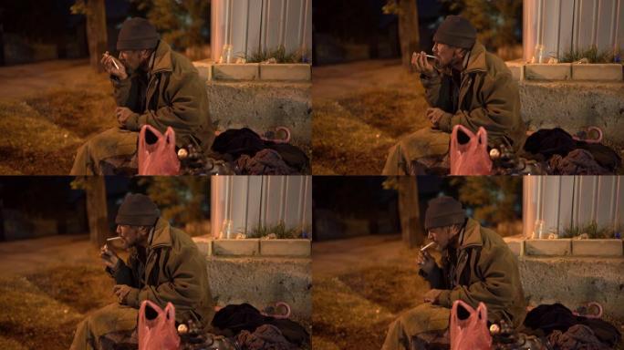孤独寒冷的无家可归的人晚上坐在街上抽烟。