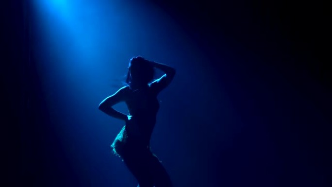 一个长发苗条的女人的剪影，她热情地跳舞拉丁美洲舞蹈的元素。一位女舞者在蓝色工作室的灯光下练习莎莎舞。