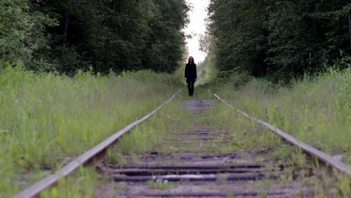 一个穿着黑色衣服的英俊年轻人在绿色森林的背景下沿着古老的铁轨行走。电影或剪辑的情节