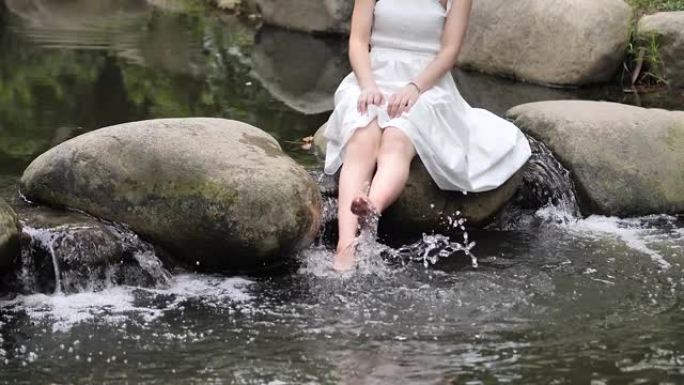 穿着白色连衣裙的赤脚年轻女子坐在大石头上，在河里玩水，快乐的女孩玩得开心，超级慢动作镜头，夏天的概念