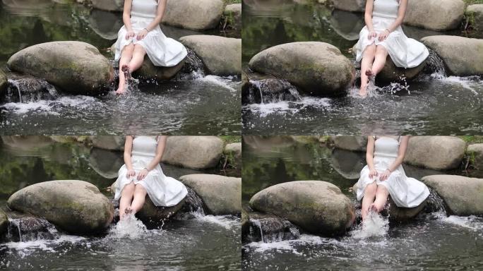 穿着白色连衣裙的赤脚年轻女子坐在大石头上，在河里玩水，快乐的女孩玩得开心，超级慢动作镜头，夏天的概念