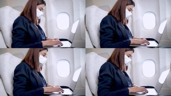 戴着口罩的亚洲妇女使用笔记本电脑，坐在窗户附近的飞机椅子上。健康保障航空旅行运输的概念新常态。